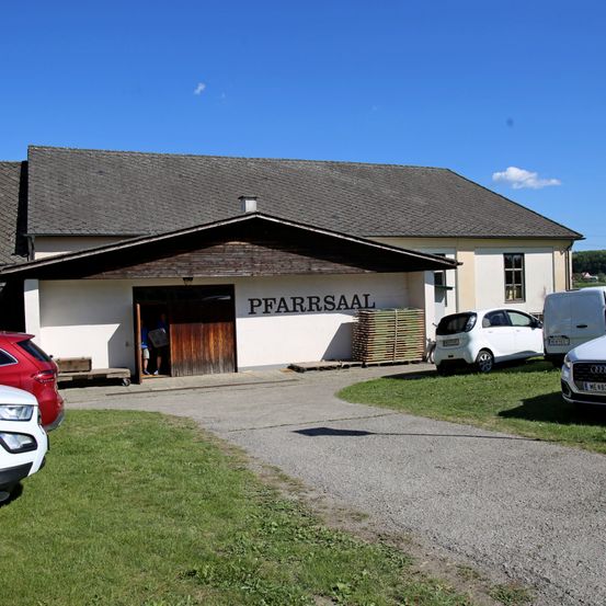 Eine Person steht am Eingang des weißen Gebäudes mit der Aufschrift 'Pfarrsaal'. Mehrere Autos sind davor geparkt, und eine Person hält ein Papier.