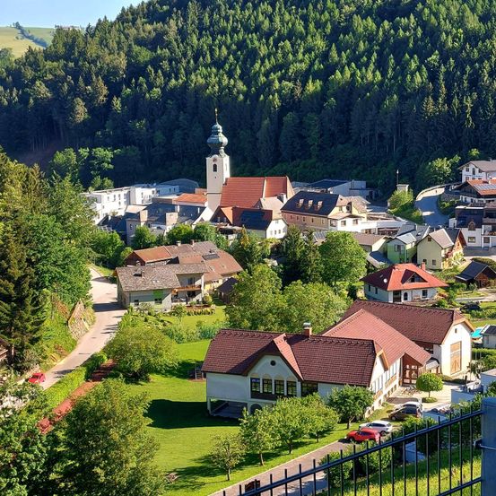 Luftaufnahme eines Dorfes in einem Tal, umgeben von Bäumen und Häusern. Eine Kirche mit einem Turm steht in der Mitte.