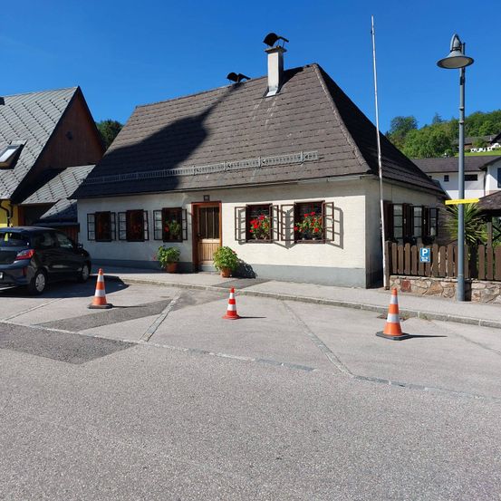 Ein kleines Haus mit einem braunen Dach und roten Blumen im Fenster. Ein Auto ist davor geparkt. Ein Zaun steht vor dem Haus. Eine Straßenlampe steht am Straßenrand.