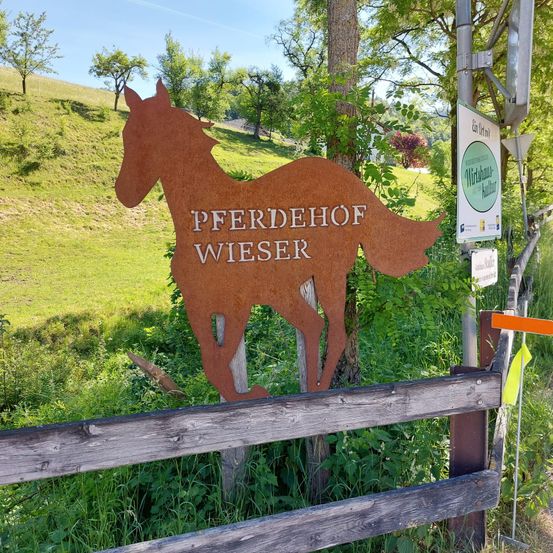 Ein großes Metallpferdeschild auf einem Bauernhof namens Pferdehof Wieser. Das Schild steht vor einer Kulisse aus grünem Gras und Bäumen.