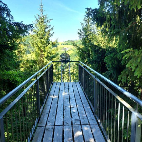 Eine hölzerne Brücke mit Metallgeländern verbindet zwei Waldbereiche und bietet einen Blick auf ein saftiges Tal.