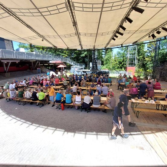 Eine große Gruppe von Menschen sitzt um Tische in einem Freiluft-Amphitheater unter einem großen Baldachin. Bäume und Treppen sind im Hintergrund zu sehen.