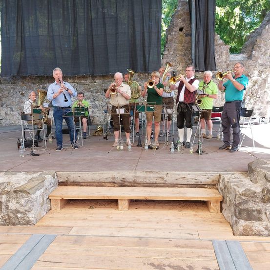 Eine Band spielt auf einer Freiluftbühne mit Steinmauern und Holzboden, gekleidet in grünen Shirts und braunen Hosen.