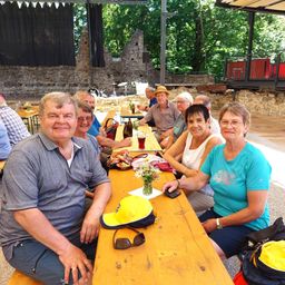 Eine Gruppe von Menschen, wahrscheinlich Freunde, sitzt auf einer Bank in einem Restaurant im Freien. Es gibt Tische mit Flaschen und einer Blumenvase. Der Mann trägt ein graues Poloshirt und Khaki-Shorts.