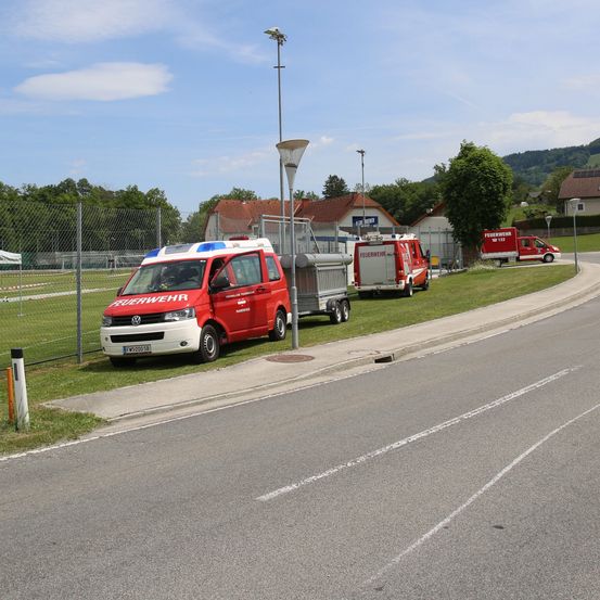 Rote Feuerwehrfahrzeuge auf einem Rasen neben einer Straße geparkt. Ein Zaun und Bäume dahinter. In der Ferne Häuser und ein Berg.