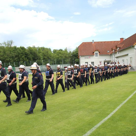 Eine Gruppe Feuerwehrleute in Uniform marschiert auf einem grünen Feld, möglicherweise während einer Trainingsübung. Das Gebäude hinter ihnen hat ein rotes Dach und weiße Wände.