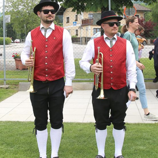 Zwei Männer in roten Westen und schwarzen Hüten stehen auf Gras und halten Trompeten. Hinter ihnen geht eine Frau an einem Zaun vorbei.