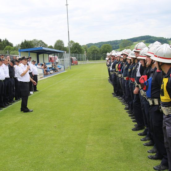 Eine Reihe von Feuerwehrleuten in Uniform steht auf einem Feld, einer von ihnen hält ein Mikrofon. Dahinter befinden sich eine Schutzhütte und ein Zaun mit geparkten Fahrzeugen.