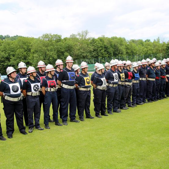 Eine Gruppe von Feuerwehrleuten in Uniform und Helmen steht in einer Reihe auf einem Feld, mit Bäumen im Hintergrund.
