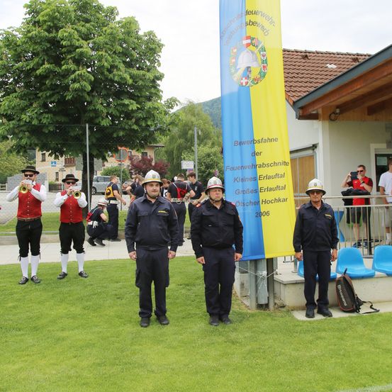 Eine Gruppe von Menschen in Uniformen steht auf einem Feld mit einer Fahne. Sie halten Trompeten und tragen Hüte. Hinter ihnen steht ein Gebäude mit einem Zaun.