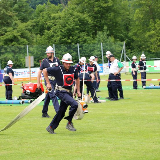 Eine Gruppe von Feuerwehrleuten trainiert auf einem Feld. Einer hält eine lange Schlauch. Die anderen stehen und beobachten.