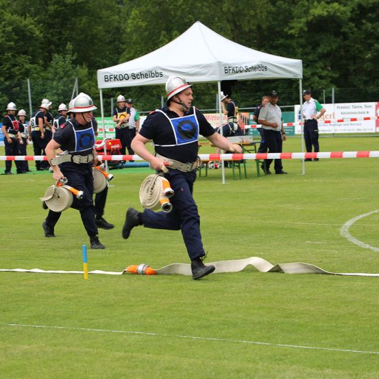 Ein Feuerwehrmann rennt mit einer Feuerwehrhose bei einem Wettbewerb auf einem Feld mit einem Zelt im Hintergrund.