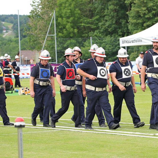 Eine Gruppe Feuerwehrleute in Uniform läuft auf einem Feld, einige tragen Helme, andere Mützen. Im Hintergrund steht ein Zelt mit der Aufschrift 'BFKDO Scheibbs' und ein Zaun.