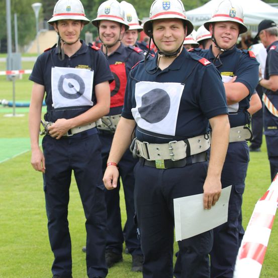 Mehrere Feuerwehrleute in Uniform stehen auf einem Feld. Der Mann vorn hält ein Papier. Im Hintergrund befinden sich Zelte und eine Menschenmenge.