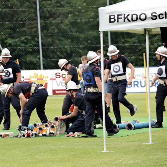 Eine Gruppe von Feuerwehrleuten bereitet sich auf einen Wettkampf unter einem Zelt vor, mit mehreren Helmen und Ausrüstungsgegenständen auf dem Boden.