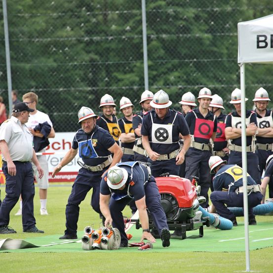 Eine Gruppe Feuerwehrleute in Uniform und Helmen arbeitet an einem roten Feuerwehrauto auf einem grünen Feld. Zuschauer beobachten von hinter einem Zaun mit einem weißen Baldachin.