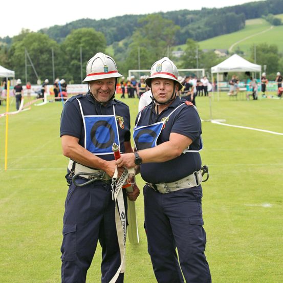 Zwei Feuerwehrleute stehen auf einem Feld mit anderen Leuten und Zelten im Hintergrund. Sie tragen beide Helme, Uniformen und Gürtel. Einer hält ein Seil.