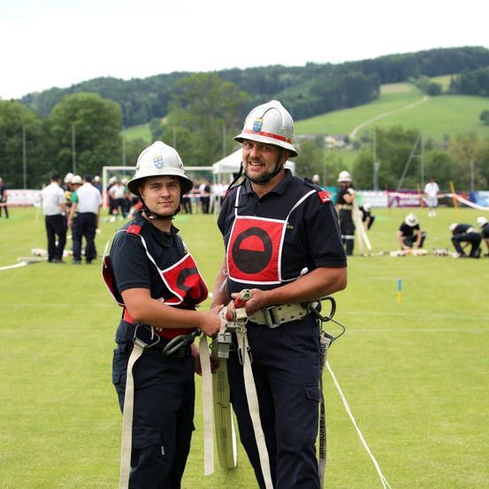 Zwei Feuerwehrleute stehen auf einem grünen Feld mit anderen Menschen im Hintergrund, einige in Helmen und einige arbeiten an Ausrüstung.