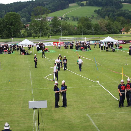 Eine Sportveranstaltung mit Teilnehmern in Uniform, Helmen und Ausrüstung auf einem Rasenfeld. Mehrere Zelte und Zuschauer sind anwesend, zusammen mit entfernten Hügeln.