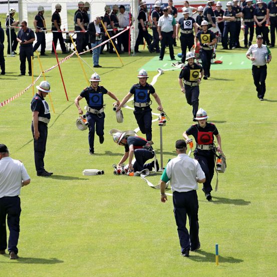 Mehrere Feuerwehrleute befinden sich auf einem Feld, einige laufen, andere stehen, mit Ausrüstung auf dem Rücken. Eine Person kniet auf dem Boden und hält ein Werkzeug.