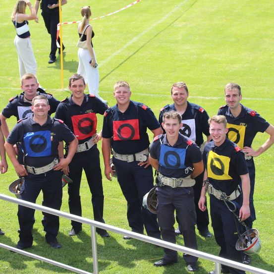Eine Gruppe lächelnder Personen in Uniform posiert auf einem Rasenfeld mit einem Metallzaun. Sie tragen farbige Westen und Helme und stehen vor einem verschwommenen Hintergrund.