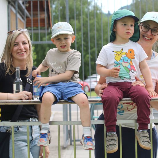 Eine vierköpfige Familie, einschließlich zweier Kinder, steht hinter einem Geländer. Die Kinder tragen Hüte und Turnschuhe. Die Frau rechts trägt eine Brille. Der Mann links hält eine Flasche. Im Hintergrund sind Bäume und ein Zaun.