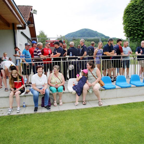 Eine Gruppe von Menschen, einschließlich Frauen und Kindern, sitzen und stehen um einen Metallzaun herum und sehen sich ein Sportereignis an. Die Menschen sind lässig gekleidet. Einige tragen Hüte und Brillen. Flaschen und Becher stehen auf dem Zaun. Auf dem Boden befinden sich Gras und Stühle. In der Ferne gibt es Bäume und Berge.