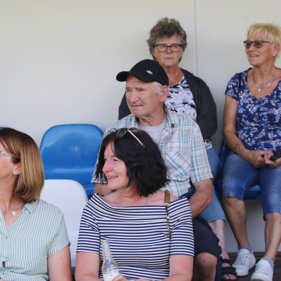 Eine Gruppe Erwachsener in lässiger Kleidung sitzt auf Stühlen. Eine Frau in einem gestreiften Shirt hält eine Flasche. Die anderen schauen in eine Richtung. Dahinter eine weiße Wand.
