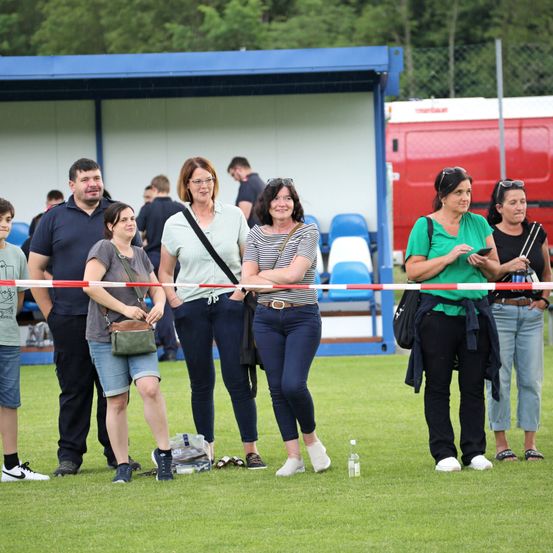 Eine Gruppe von Menschen steht hinter einem rot-weißen Band auf einem Rasenfeld, wahrscheinlich in Erwartung einer Veranstaltung. Ein roter Van ist dahinter geparkt, und ein blaues Gebäude ist sichtbar.