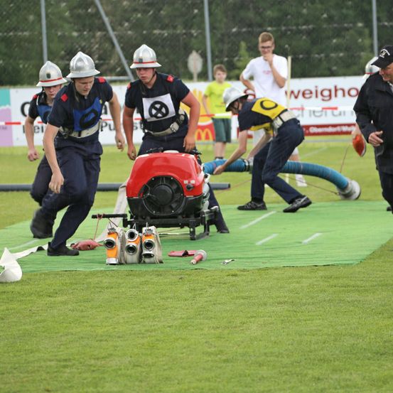 Feuerwehrleute in Uniformen üben auf einem grünen Feld. Ein Feuerwehrmann benutzt einen Schlauch, um ein Feuer zu löschen. Ein Polizist steht auf der rechten Seite.