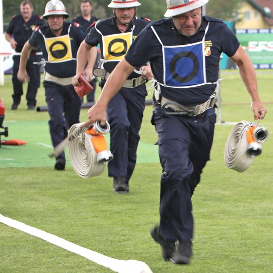 Mehrere Feuerwehrleute mit Helmen laufen auf einem Feld und tragen einen Feuerwehrschlauch. Der Mann vorne hält einen Schlauch. Sie nehmen wahrscheinlich an einem Feuerwehrwettbewerb teil.
