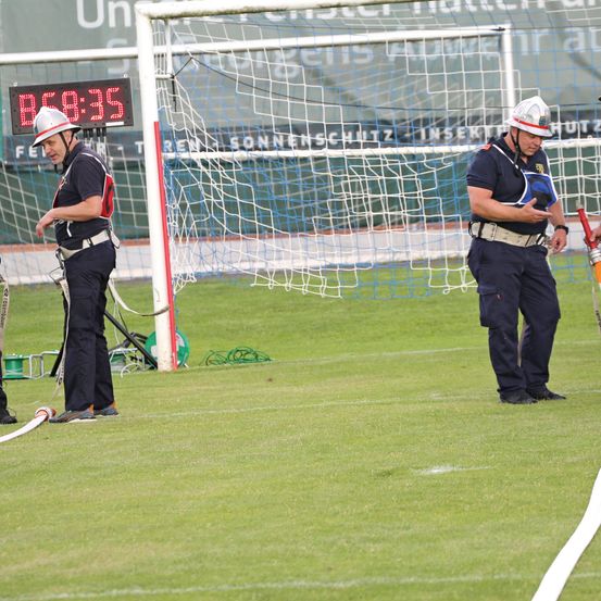 Zwei Männer in Sicherheitsausrüstung, Helmen und Gürteln stehen auf einem Feld in der Nähe eines Fußballtors mit einer digitalen Uhr, die 88:35 anzeigt.