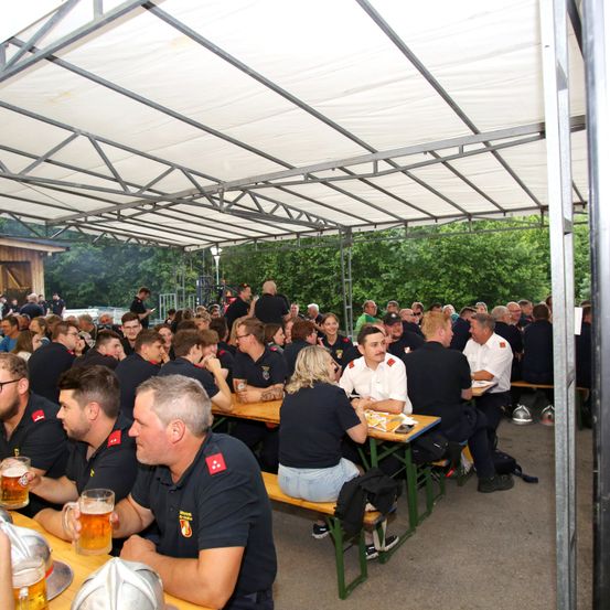Eine große Gruppe von Feuerwehrleuten hat sich unter einem Zeltdach zum Essen versammelt. Einige sitzen an Tischen, andere stehen und unterhalten sich. Biergläser und Helme sind vorhanden.