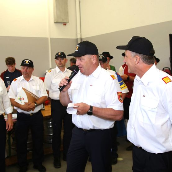 Eine Gruppe von Feuerwehrleuten in Uniform. Ein Mann spricht in ein Mikrofon, während andere aufmerksam zuhören. Sie befinden sich in einem Raum mit grauen Wänden und einem Tisch im Hintergrund.
