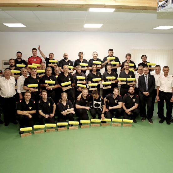 Eine Gruppe Feuerwehrleute, gekleidet in schwarze Uniformen, posiert für ein Foto und hält gelbe Objekte. Sie befinden sich in einem Raum mit weißen Wänden und grünem Boden, umgeben von anderen Menschen.
