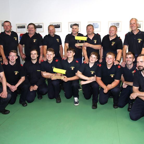 Eine Gruppe Feuerwehrleute, in schwarzen Uniformen gekleidet, posiert für ein Foto. Einige halten Trophäen. Sie stehen auf einer grünen Oberfläche mit gerahmten Bildern an einer weißen Wand hinter ihnen.