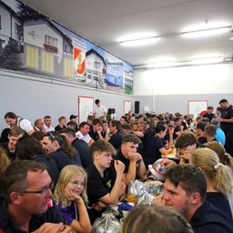 Eine große Gruppe von Menschen in Uniform sitzt an einem langen Tisch in einem Raum. Der Raum hat hohe Decken und große Wandgemälde. Die Personen scheinen in Gespräche vertieft zu sein und einige halten Helme.