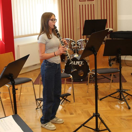 Ein Mädchen steht in einem Musikstudio und spielt Klarinette, mit einem Schlagzeug und Stühlen im Hintergrund.