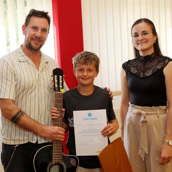 Ein junger Junge hält ein Zertifikat und eine Gitarre in der Hand und steht zwischen einem Mann und einer Frau, alle lächeln für die Kamera. Der Junge trägt ein schwarzes Shirt und der Mann ein gestreiftes Hemd.