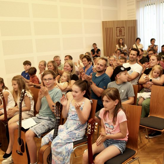 Eine Gruppe von Kindern und Erwachsenen klatscht in einem Saal mit Holzstühlen. Einige halten Gitarren, während andere ihre Hände klatschen. Der Raum hat eine Holzwand und weiße Jalousien.