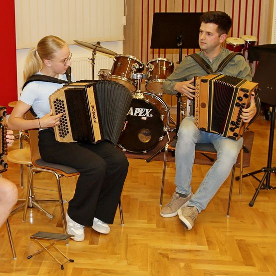 Zwei Personen sitzen auf Stühlen und spielen Akkordeons. Dahinter steht ein Schlagzeug. Die Person auf der linken Seite trägt eine Brille und Turnschuhe. Die Person auf der rechten Seite trägt eine Uhr und Turnschuhe.