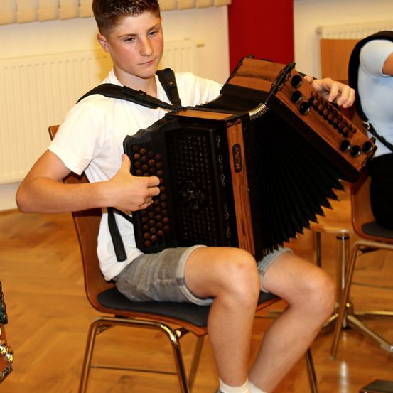 Ein junger Junge sitzt auf einem Stuhl und spielt Akkordeon. Er ist in einem weißen T-Shirt und Shorts gekleidet. Er befindet sich in einem Raum mit Holzboden und weißen Wänden.