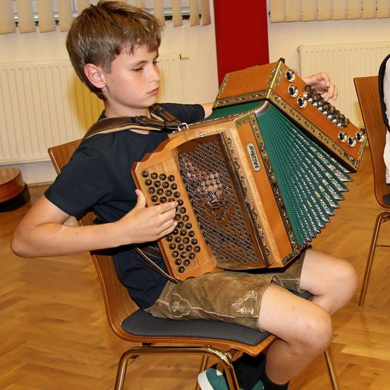 Ein junger Junge in einem schwarzen T-Shirt und braunen Shorts spielt Akkordeon, während er auf einem Stuhl sitzt.