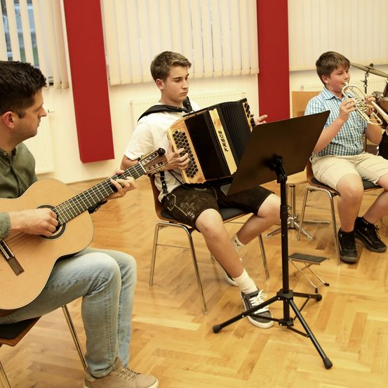Drei Jungen sitzen auf Stühlen und spielen Instrumente. Einer spielt Gitarre, ein anderer spielt Trompete und der Mittlere spielt Akkordeon. Sie sind in einem Raum mit Holzboden und einer roten Wand.