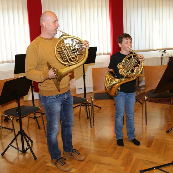 Ein Mann und ein Junge stehen in einem Raum und halten jeweils ein französisches Horn aus Messing. Sie befinden sich in einem Musikstudio.