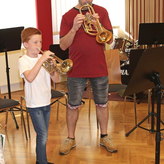 Ein junger Junge spielt Trompete, während ein Mann neben ihm steht, beide in einem Raum mit Musikinstrumenten und Notenblättern.