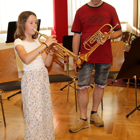 Ein Mädchen in einem Blumenrock spielt eine Trompete in einem Musikraum, wobei ein Mann in einem roten T-Shirt ebenfalls eine Trompete hinter ihr spielt.