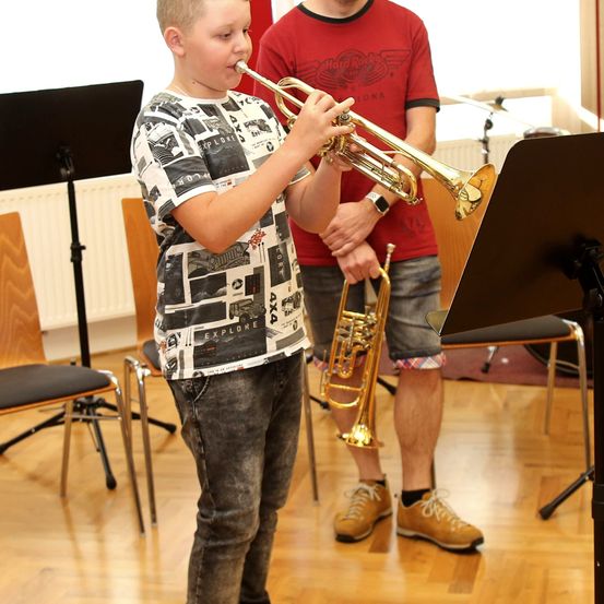 Ein junger Junge spielt Trompete in einem Musikzimmer, während ein Lehrer in der Nähe steht und ebenfalls eine Trompete hält.