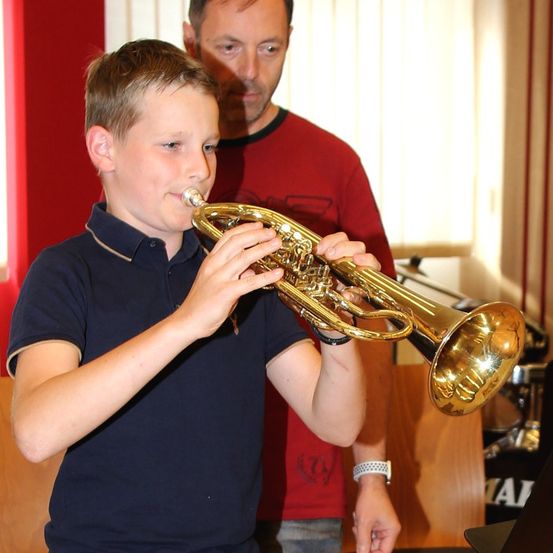 Ein junger Junge spielt eine goldene Trompete, während ein Mann in einem roten T-Shirt zusieht. Der Junge trägt ein marineblaues Poloshirt und sieht konzentriert aus. Dahinter befindet sich eine rote Wand und ein weißer Vorhang.