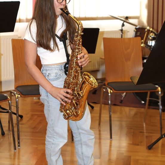 Eine junge Frau steht in einem Raum und spielt eine Saxophon, während Stühle und ein Schlagzeug in der Nähe sind.